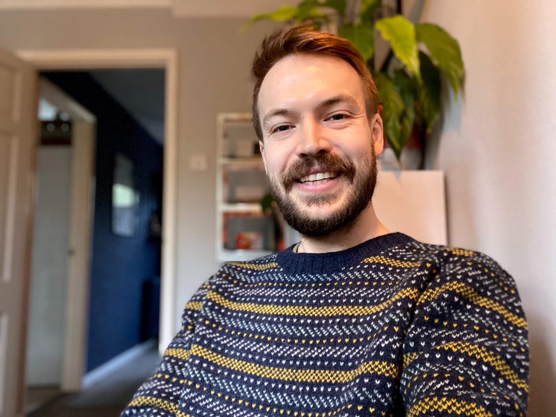 Smiling man with beard (Barnaby Mollett) in patterned sweater, standing indoors with plants and shelves blurred behind him.