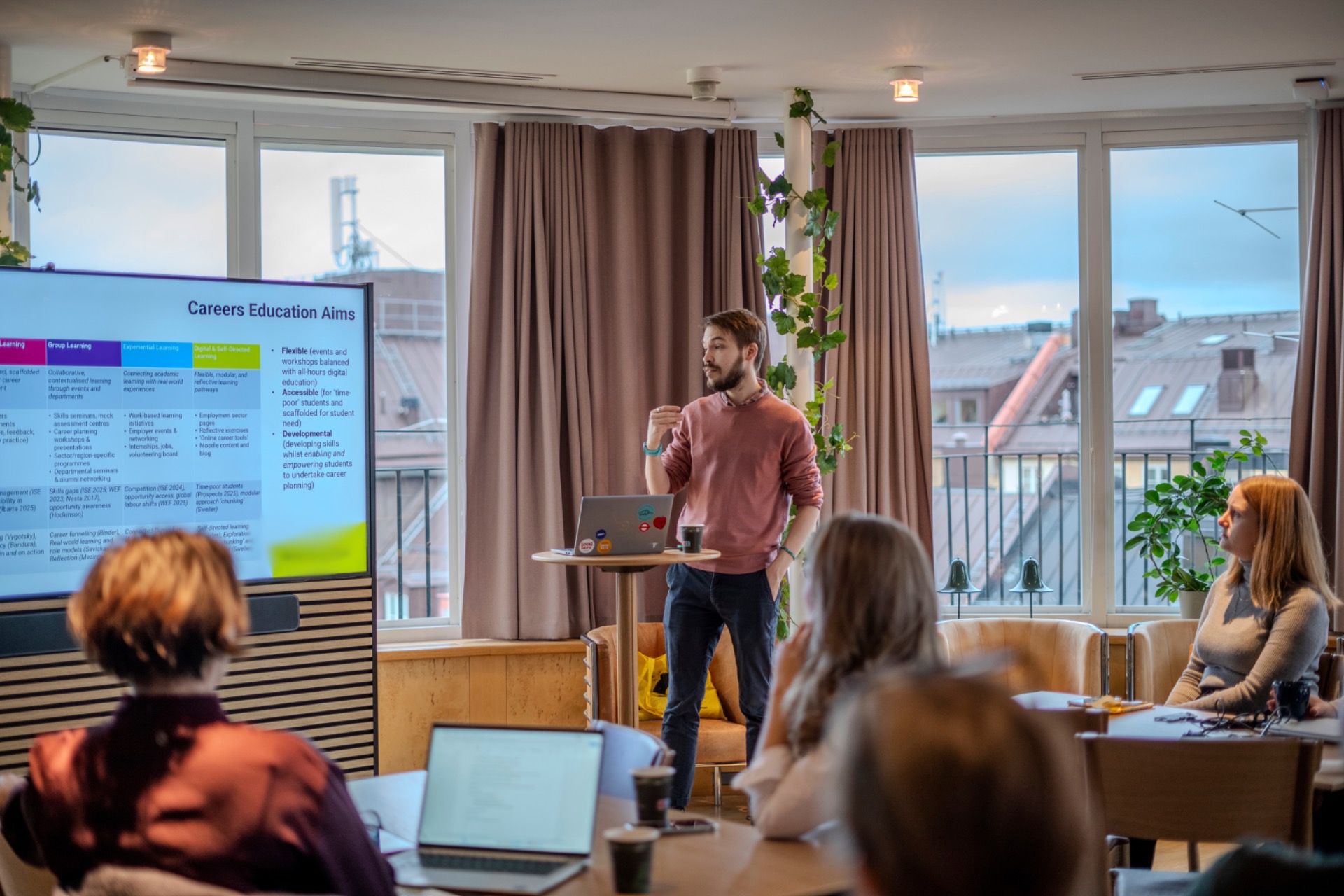 Presenter (Barnaby Mollett) stands by laptop during meeting, audience seated, next to big TV screen with a slide that reads “Careers Education Aims” in bright conference room.