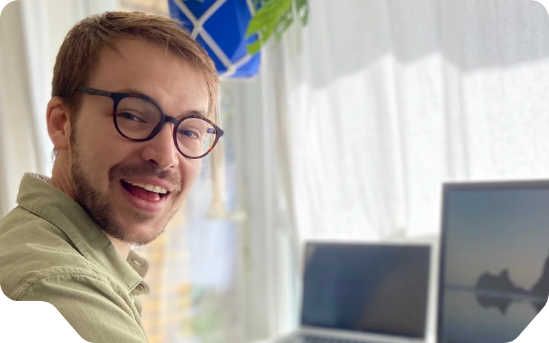 Smiling man (Barnaby Mollett) with glasses working at a desk, typing on a keyboard beside two screens: a monitor and a laptop.