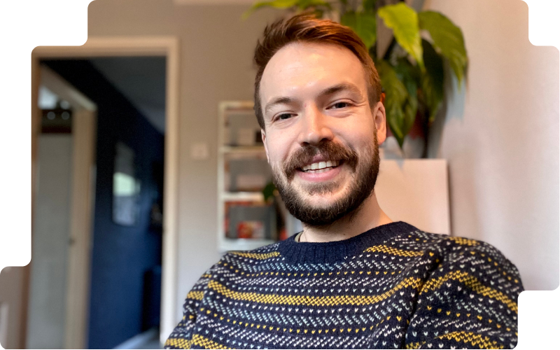 Smiling man with beard (Barnaby Mollett) in patterned sweater, standing indoors with plants and shelves blurred behind him.