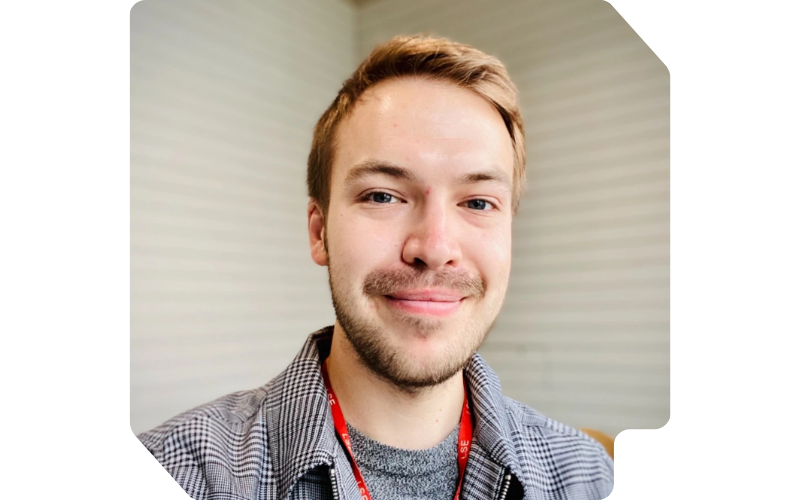 Smiling man (Barnaby Mollett) with short blond hair wearing a checkered shirt and red lanyard indoors.