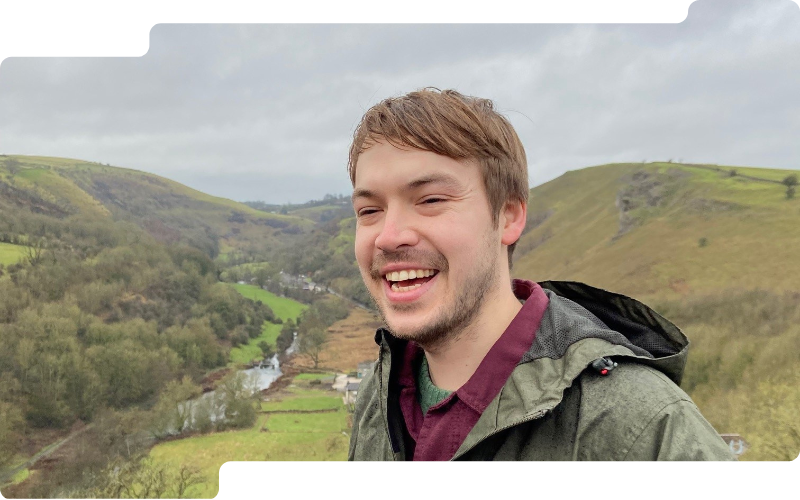 Smiling man (Barnaby Mollett) in outdoor jacket taking selfie overlooking green valley hills under cloudy sky.