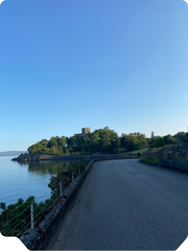 Quiet lakeside road curves past trees toward a small stone tower under a clear blue sky.